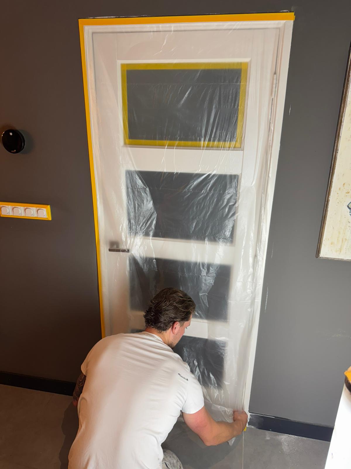 Schilder zet MOOZERS maskeerfolie vast op een witte paneeldeur die is afgeplakt met gele Washi tape